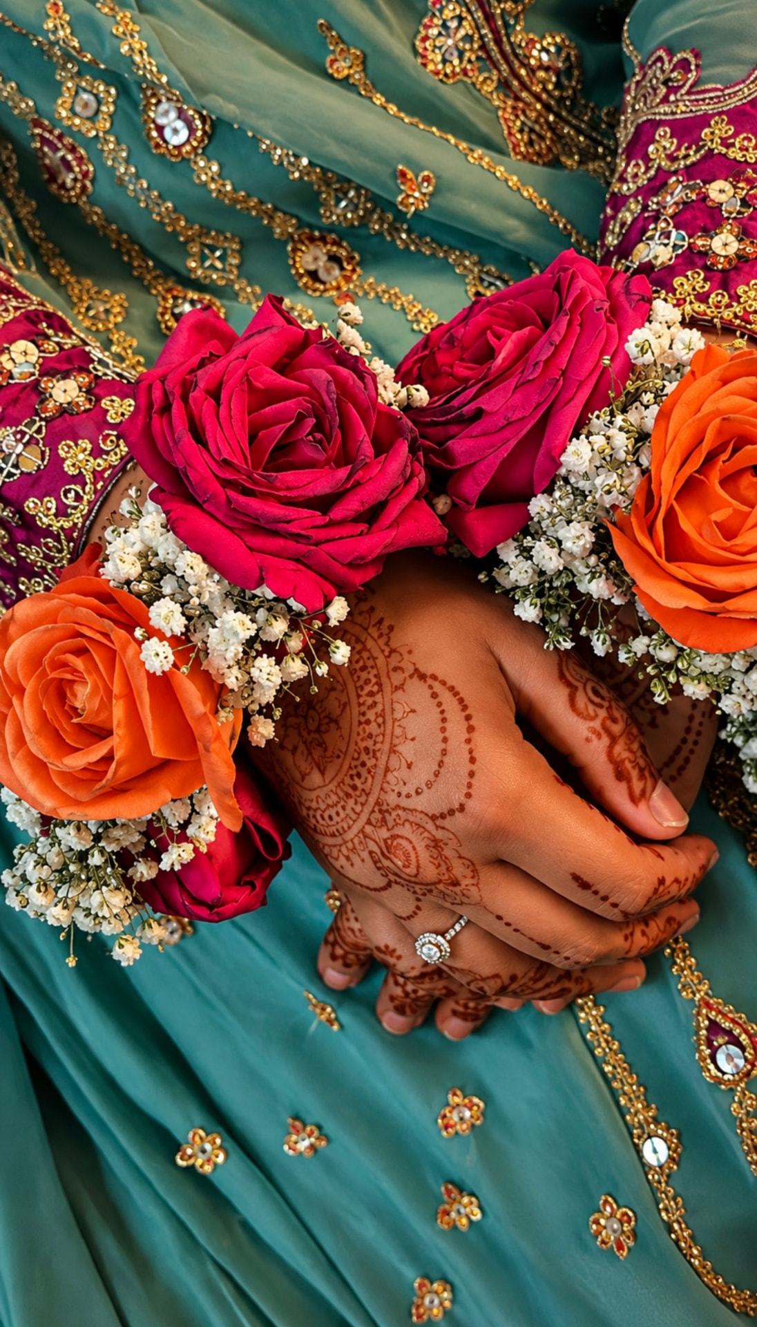 Bridal hands with henna resting on a teal and gold outfit, wearing gajras of red and orange roses with baby's breath